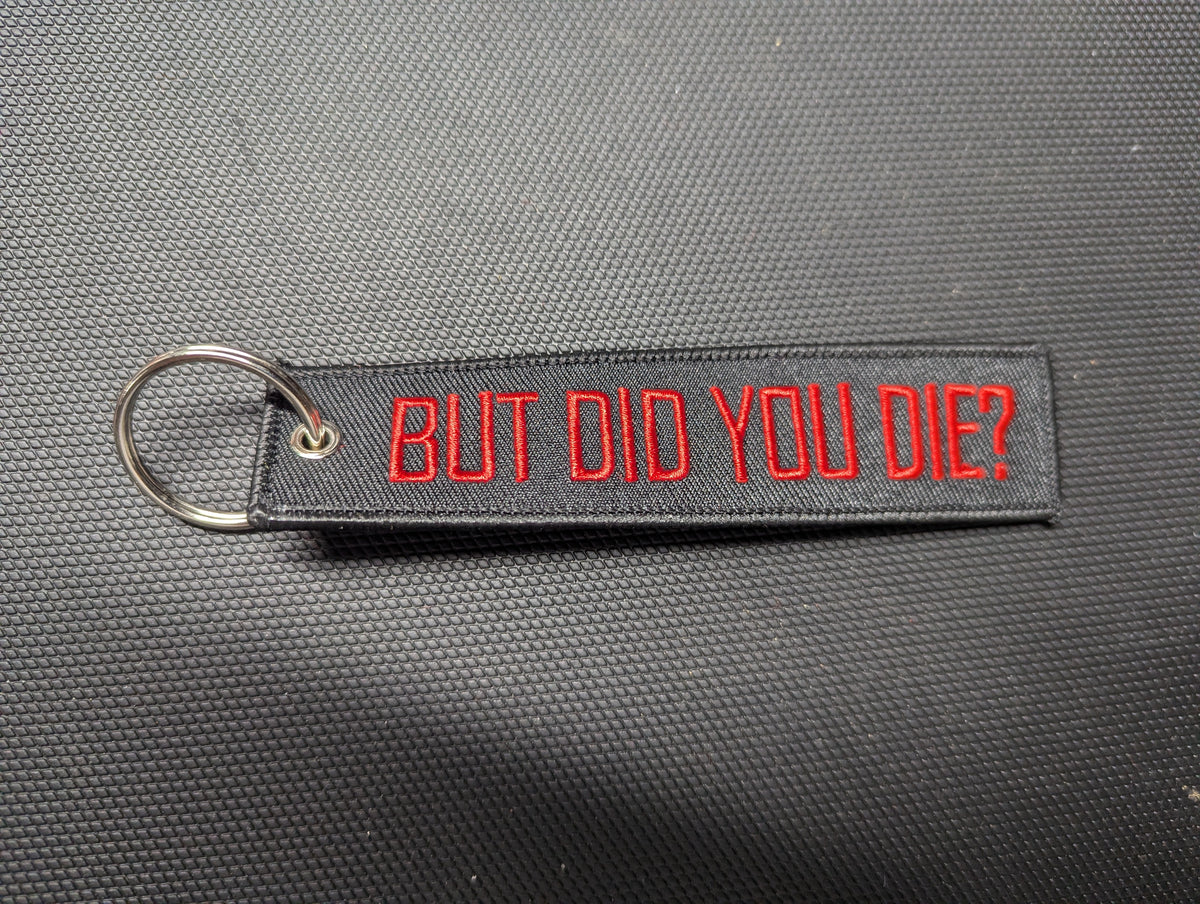Black fabric keychain embroidered in red with “BUT DID YOU DIE?” and silver-tone split ring, upright